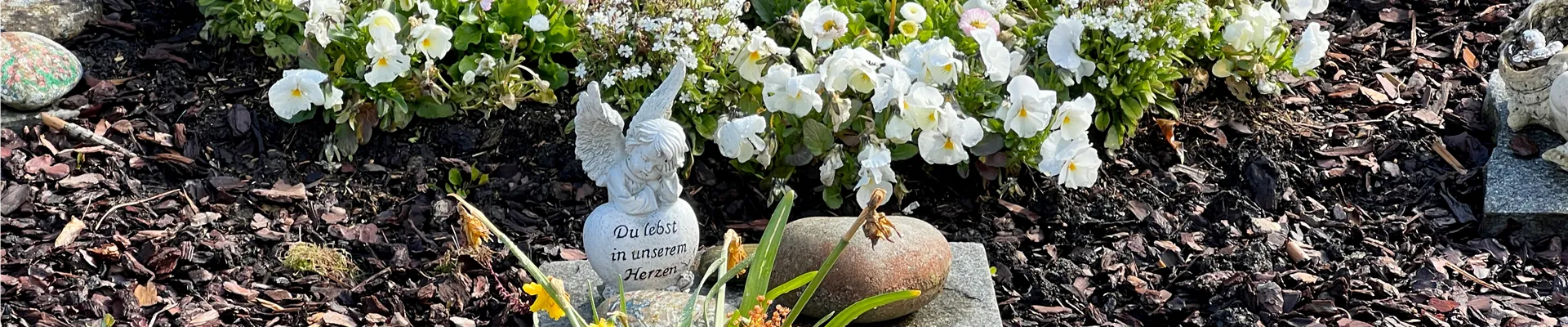 Blumenbeet mit weißen Blumen und einem kleinen Steinschild mit der Aufschrift 'Du lebst in unseren Herzen'